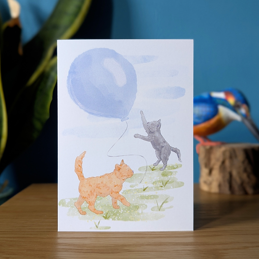 grey kitten birthday card with ginger cat trying to catch a big blue balloon
